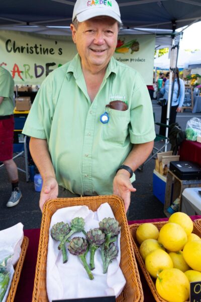 Man at a farmers market stall proudly displaying fresh artichokes in a wicker basket, with lemons visible in another basket, promoting local heirloom produce from Christine's Garden.