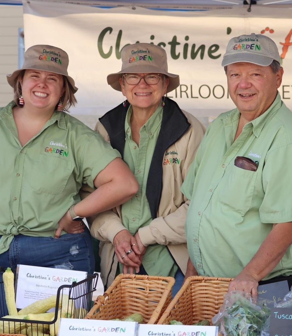 Christine's+Garden,+California+-+29D8FE43-5133-4CF3-8986-AB30C6293432 Three smiling vendors at Christine's Garden farmer's market stand, wearing matching green shirts and hats. The table features baskets of fresh produce and a sign displaying the garden's name.