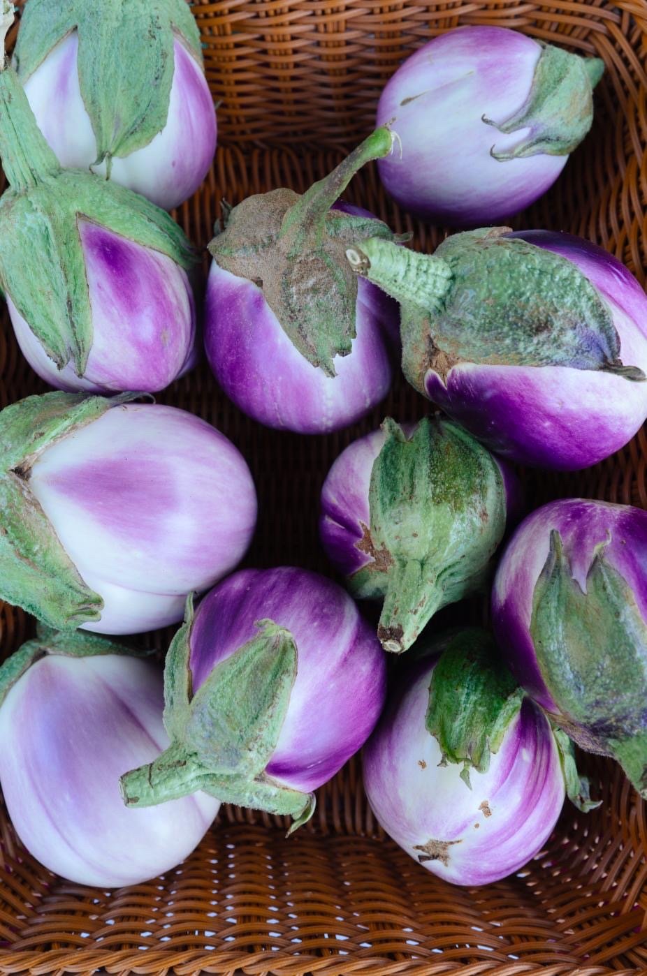 Christine's+Garden,+California+-+4F6627EC-E6AF-463E-BAF7-6E9DD56CB59E Fresh purple eggplants arranged in a wicker basket, showcasing their glossy skin and green tops, ideal for healthy cooking and recipes.