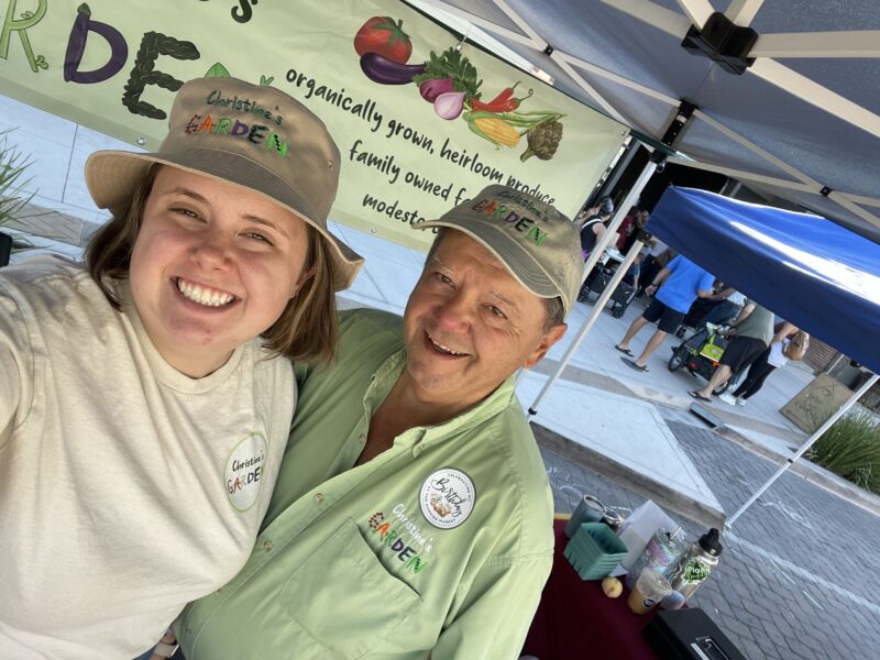 Smiling team members at Christine's Garden, a family-owned business offering organically grown heirloom produce, pose in front of their market stall with a colorful banner. The scene captures a vibrant farmers market atmosphere with customers in the background.