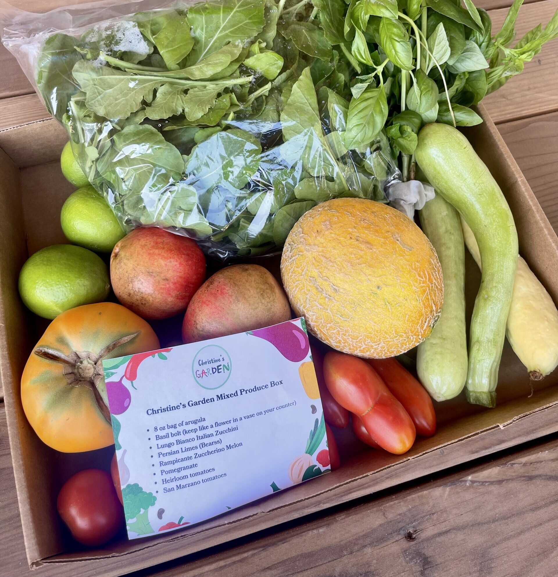 A cardboard box filled with fresh produce, including a variety of vegetables and fruits such as basil, arugula, zucchini, melons, apples, and tomatoes, alongside a label detailing the contents of Christine's Garden Mixed Produce Box.