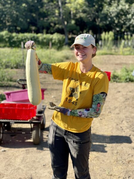 A smiling woman in a yellow t-shirt holds a large zucchini in a community garden, surrounded by lush greenery and gardening equipment. Her long-sleeved gloves feature colorful vegetable patterns, highlighting her passion for gardening and sustainable agriculture.