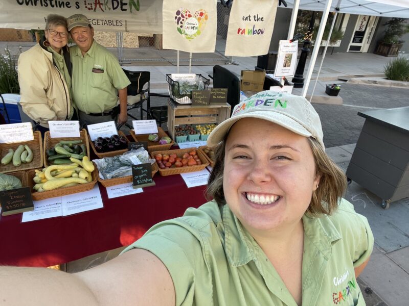 A smiling young woman takes a selfie at a farmer's market booth for Christine's Garden, featuring a variety of fresh organic vegetables, including zucchini, kale, and tomatoes. Two vendors, an older woman and a man, stand behind the table, promoting their produce. The booth is decorated with signs encouraging customers to 